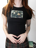 A person wearing a black short-sleeve graphic T-shirt with a rectangular printed photo collage on the chest and a plaid skirt, standing indoors against a light wall.