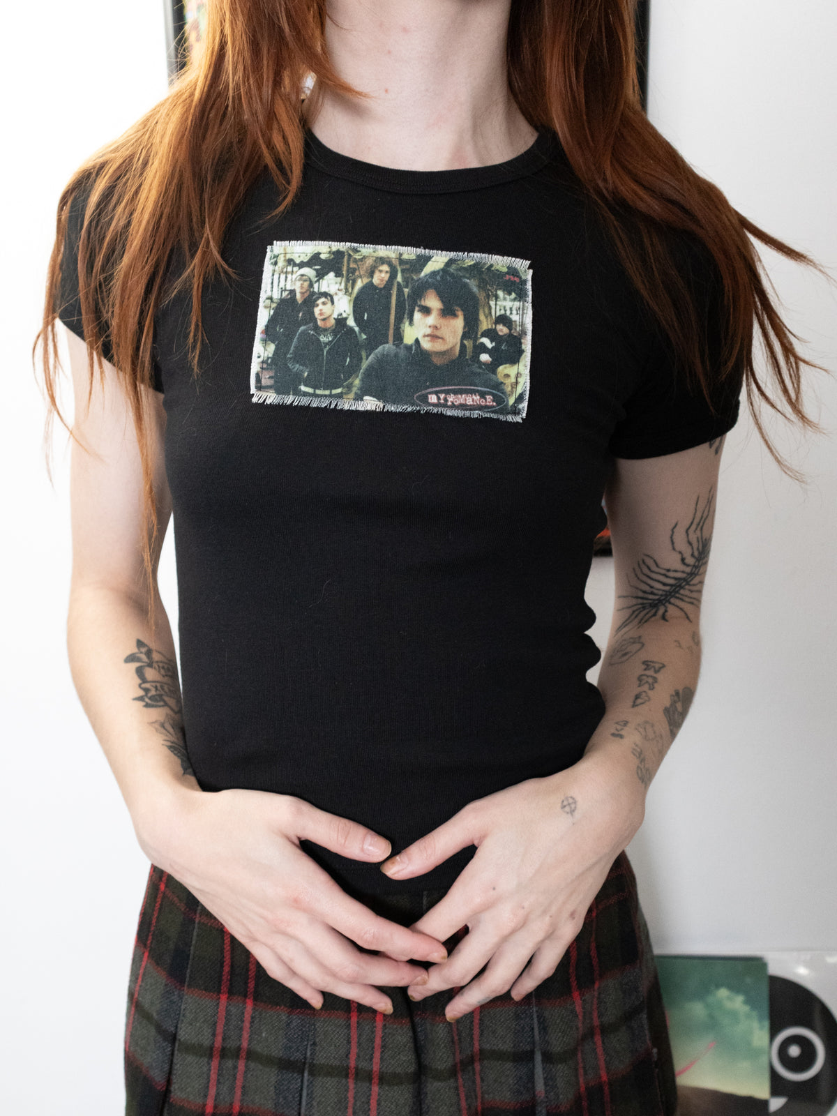 A person wearing a black short-sleeve graphic T-shirt with a rectangular printed photo collage on the chest and a plaid skirt, standing indoors against a light wall.