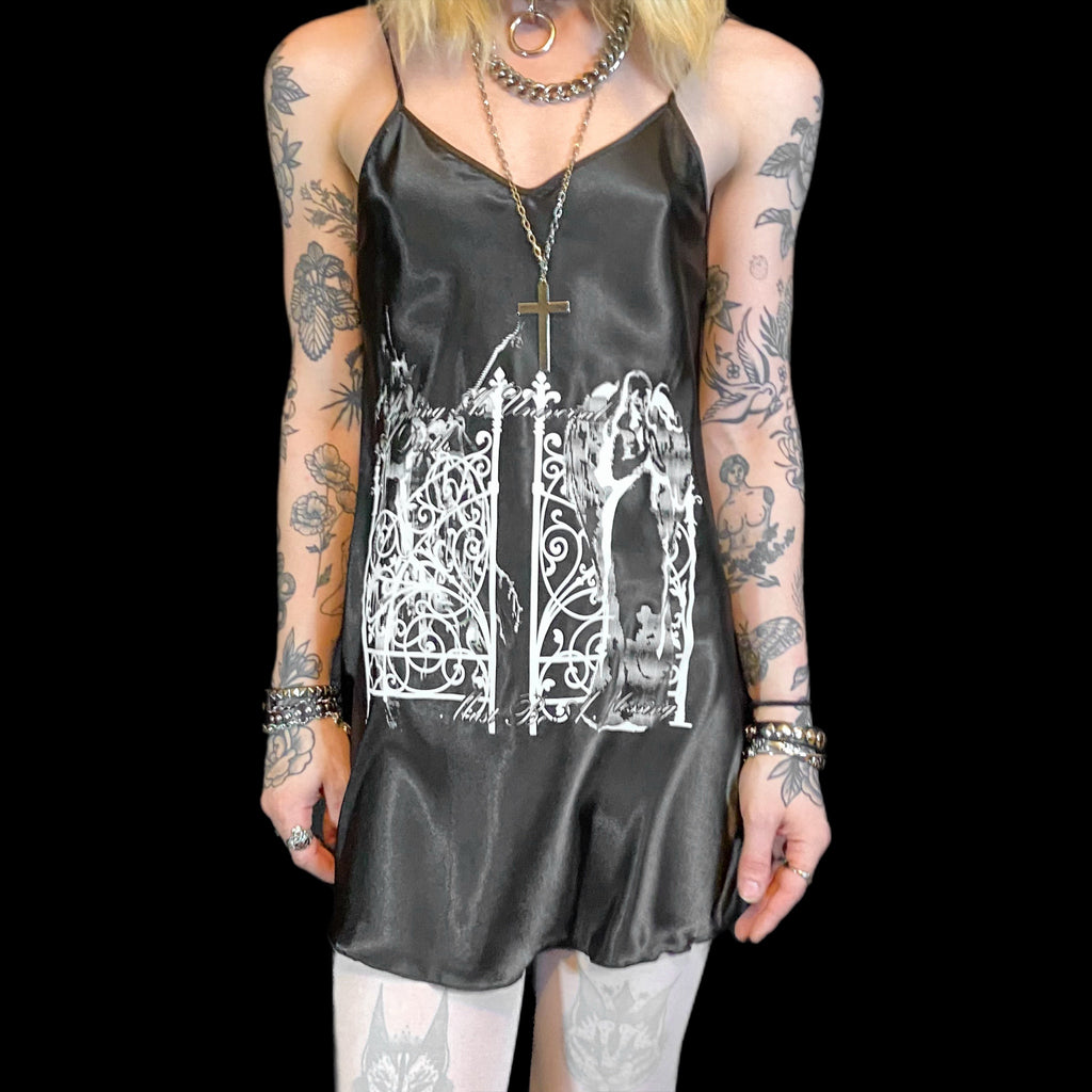 A person wearing a black slip dress with a screen printed design of an angel and text, paired with silver accessories and a cross necklace.