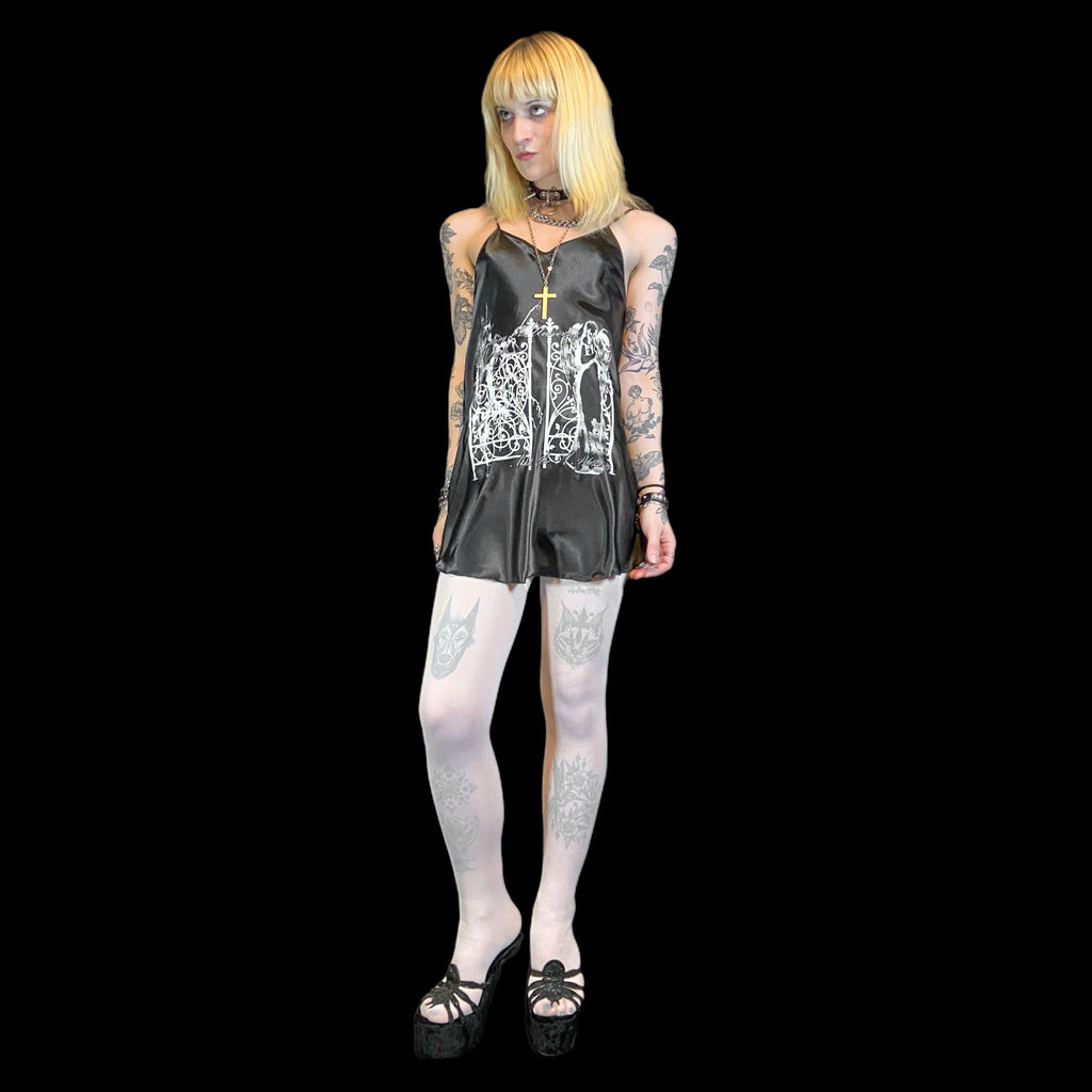 Person wearing a black slip dress with white graphic design, fishnet stockings, and black shoes on a black background.