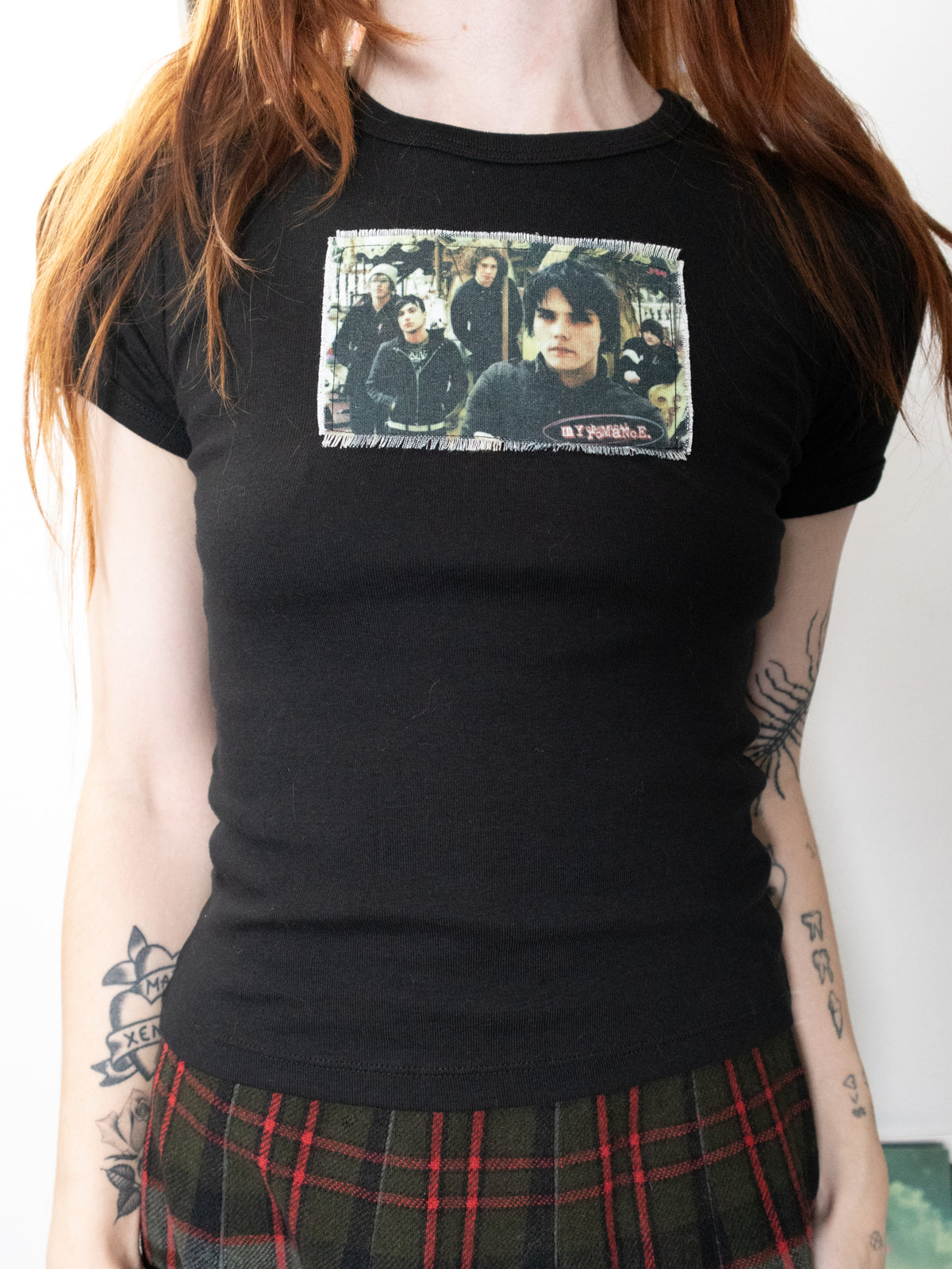 A person wearing a black short-sleeve graphic T-shirt with a rectangular band print on the chest, paired with a green and red plaid skirt. The shirt features a photo print of the band my chemical romance