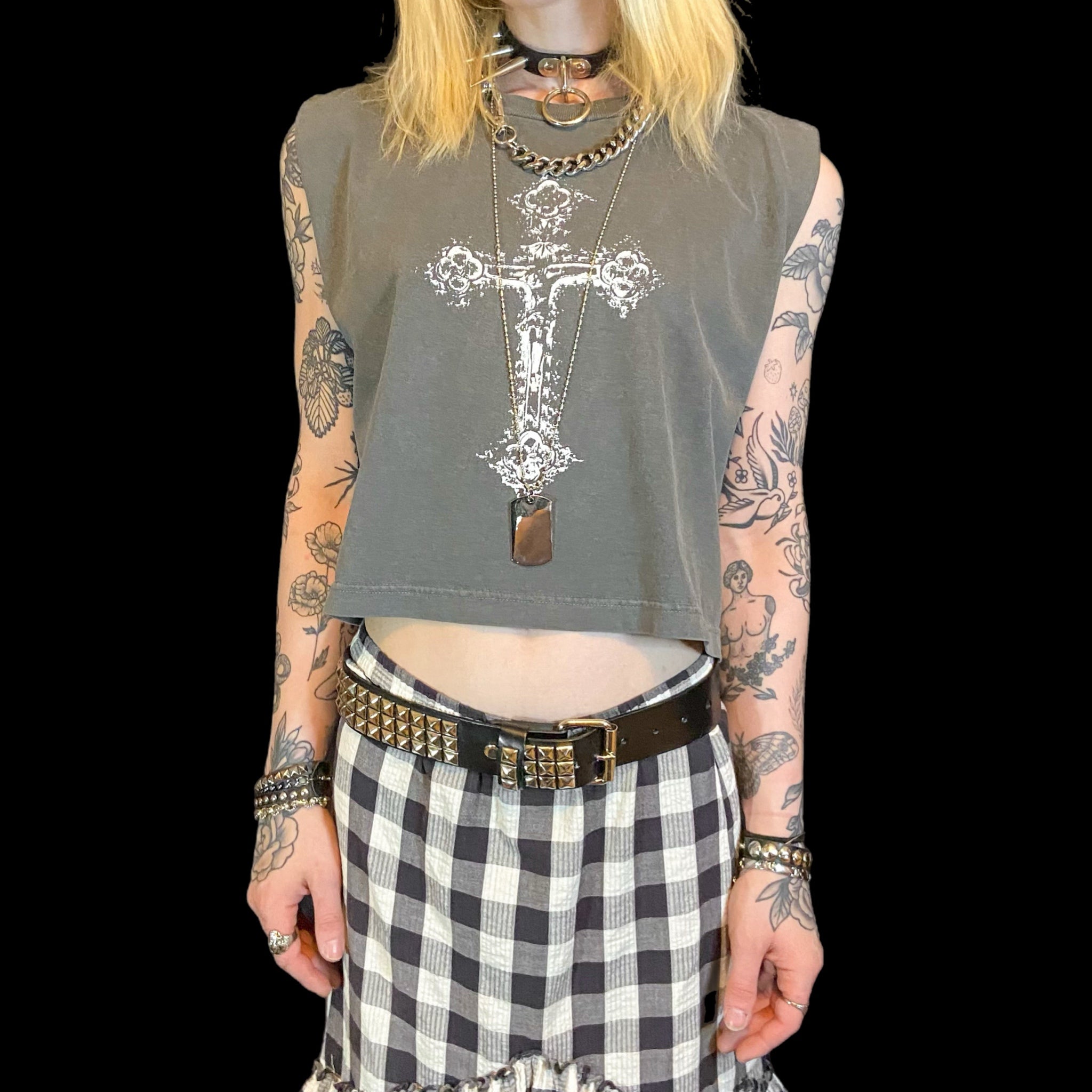 A person wearing a gray muscle tank top with a white hand-printed design that resembles a cross and has the brand name 'trashgirl'. The person is also wearing a plaid skirt and has multiple necklaces.
