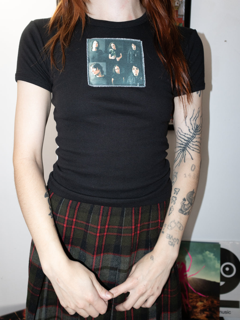 A person wearing a fitted black short-sleeve t-shirt with a rectangular band graphic on the chest, paired with a dark plaid skirt.