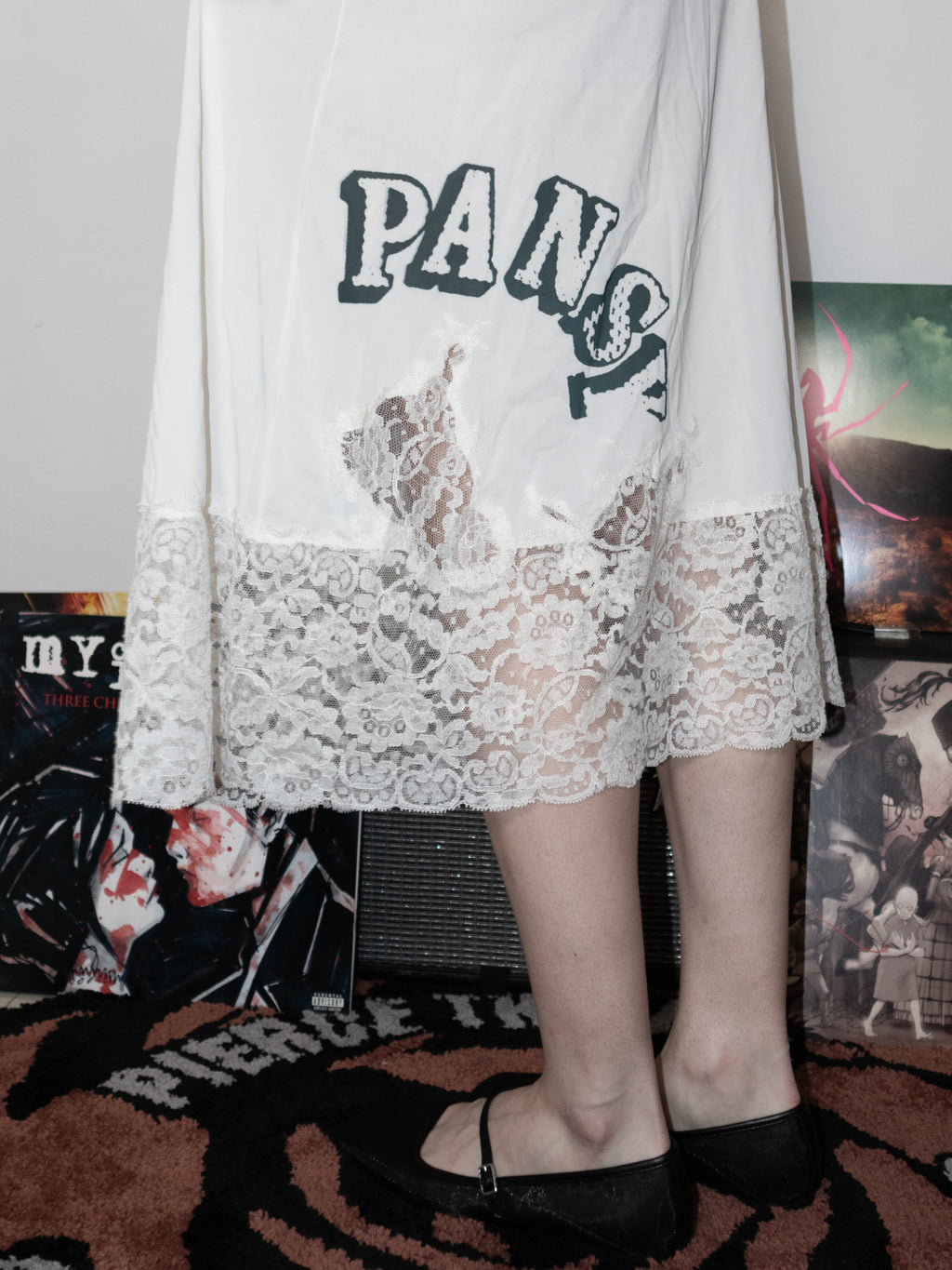 A person wearing a white sheer long-sleeve top or robe with a large black 'Pansy!' graphic and a lace hem, paired with black flats, standing in a room with posters and a speaker in the background.