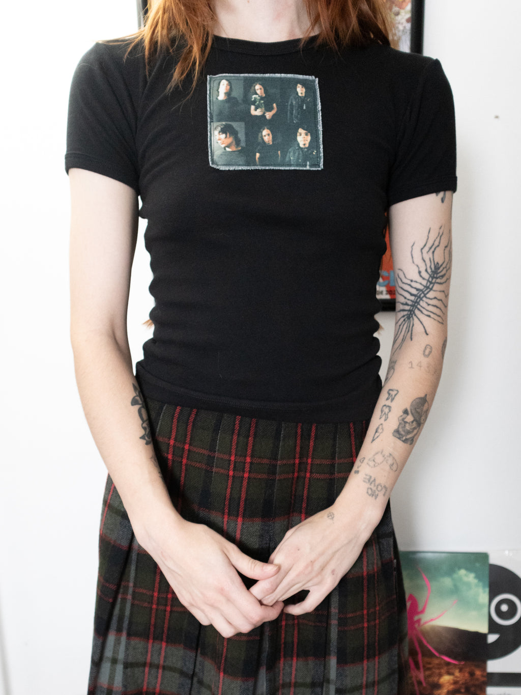 A person wearing a fitted black short-sleeve T-shirt with a rectangular band graphic on the chest, paired with a dark plaid skirt, standing indoors against a light wall.