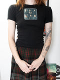 A person wearing a fitted black short-sleeve T-shirt with a rectangular band graphic on the chest, paired with a dark plaid skirt, standing indoors against a light wall.