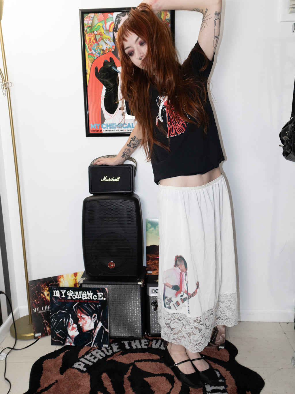 A woman with long red hair poses in a room wearing a black graphic crop top, a long white skirt with lace hem, and black strappy heels. She stands next to a Marshall speaker and a speaker stack on a patterned rug, with posters on the wall behind her.