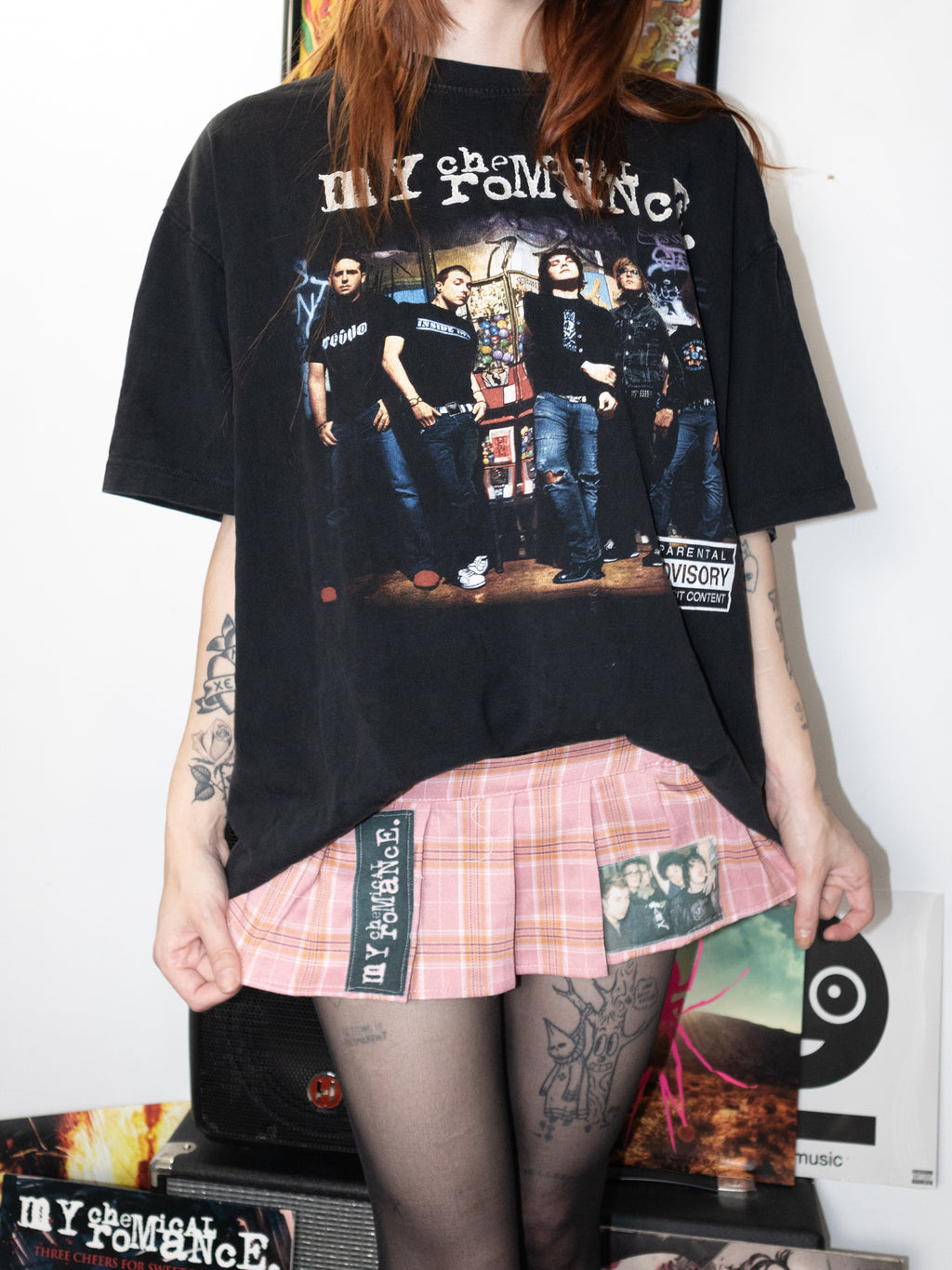 A person wearing an oversized black graphic T-shirt with a band photo and text, paired with a pink plaid pleated skirt and black tights, standing indoors against a white wall.