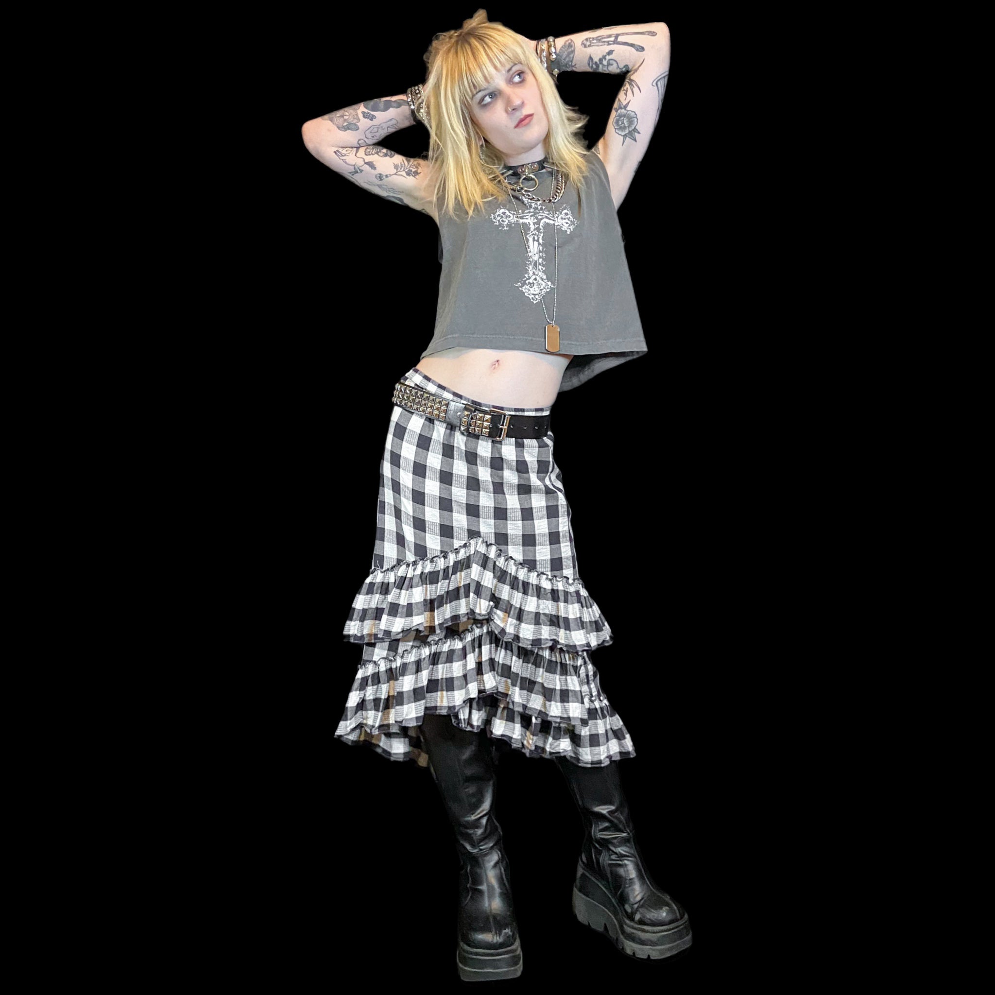 Person wearing a gray sleeveless top with a cross design, black and white checkered skirt, and black boots on a black background.