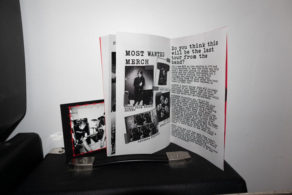 An open printed booklet or magazine spread displayed on a black stand, with a small black-and-white photo print leaning beside it against a plain light wall.