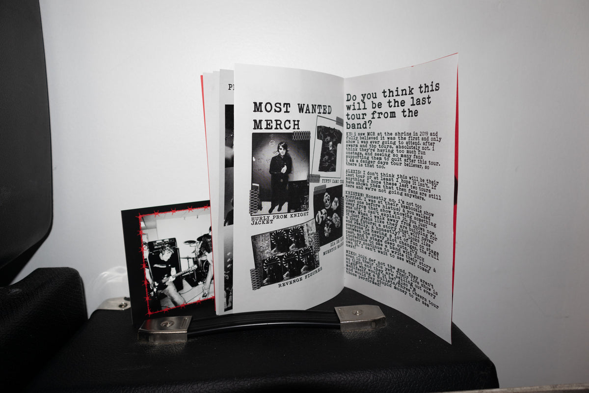 An open printed booklet or magazine spread displayed on a black stand, with a small black-and-white photo print leaning beside it against a plain light wall.
